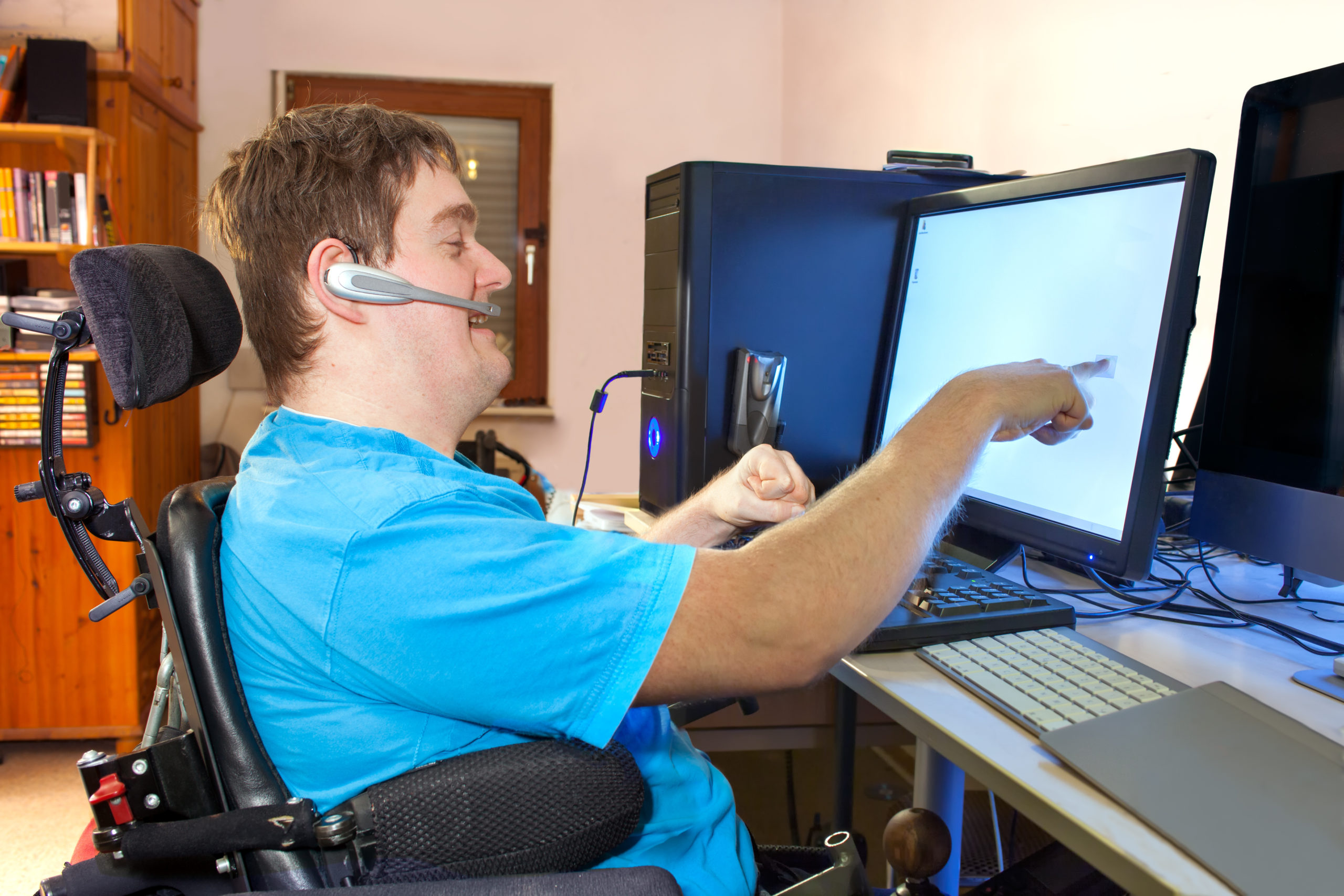 Young disabled man with cerebral palsy sitting in a powerchair using a computer with a wireless headset reaching out to touch the computer screen
